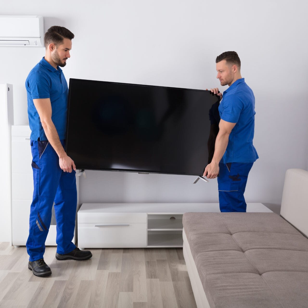 Two Young Male Movers In Uniform Placing Television In Living Room